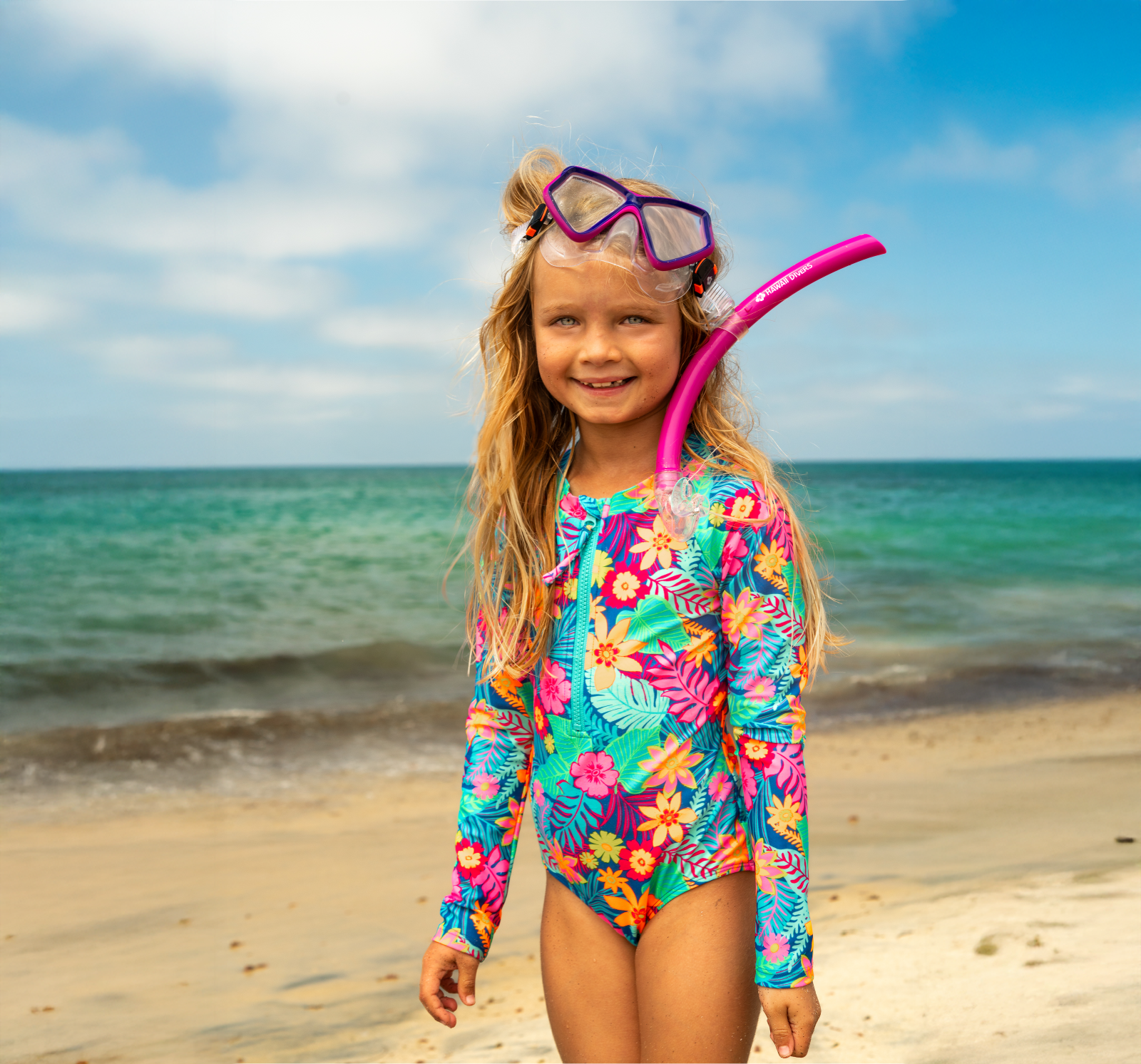Child on a beach wearing a colorful swimsuit and snorkeling gear with a blue sky and ocean background.