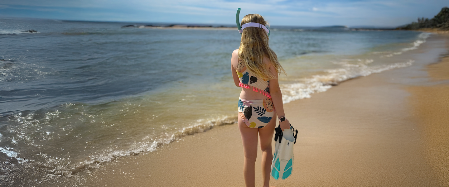Woman standing on a beach holding snorkeling gear with ocean waves in the background